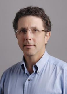 Image is head shot of Jon Amster, caucasian male with curly hair and glasses, wearing a blue shirt