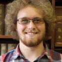 Headshot of Jason Colley, white male with curly blond hair, beard, and glasses; background of books in a library